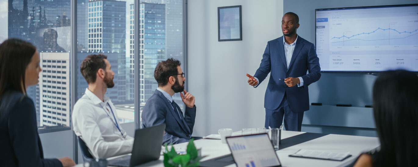 Diverse Office Conference Room Meeting: Charismatic Black Digital Entrepreneur Presents e-Commerce Fintech Growth Statistics to a Group of Investors. Wall TV with Big Data Analysis, Infographics